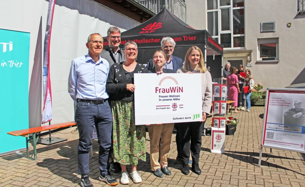 Drei Frauen und drei Männer stehen in einem sonnigen Hinterhof und halten lächelnd ein Schild mit der Aufschrift "FrauWIN" in die Kamera.