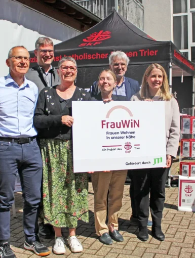 Drei Frauen und drei Männer stehen in einem sonnigen Hinterhof und halten lächelnd ein Schild mit der Aufschrift "FrauWIN" in die Kamera.